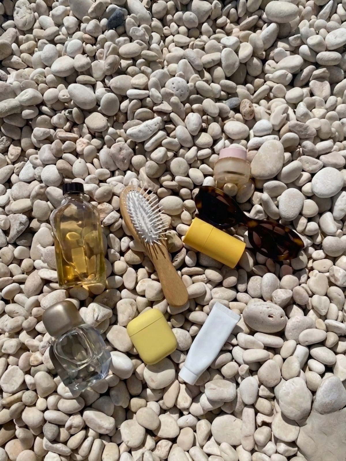 Närbild på personliga accessoarer som glasögon och hudvårdsprodukter placerade på en strand med ljusa, runda stenar.