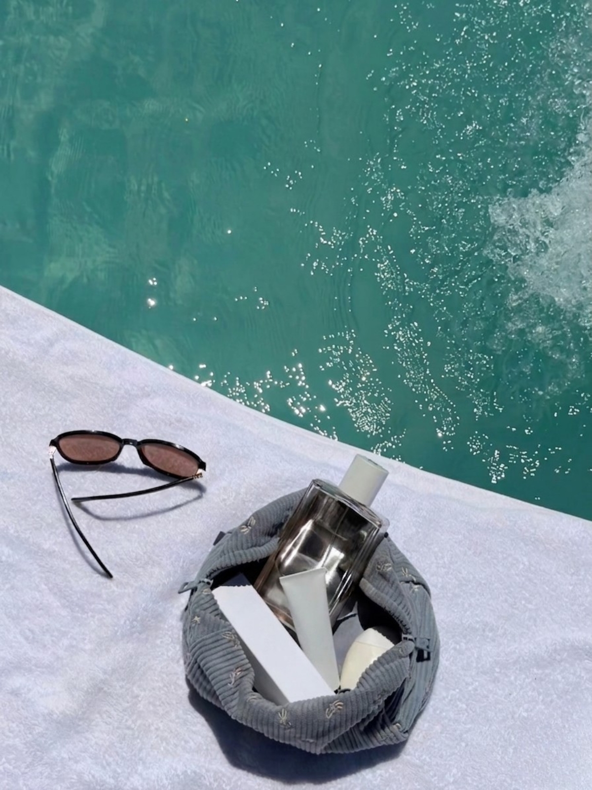 Somrig scen vid en pool med solglasögon och en necessär med hudvårdsprodukter på en vit handduk.